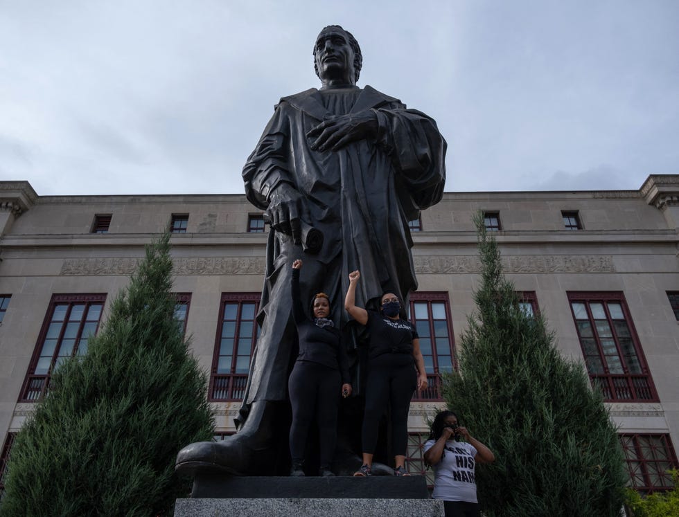 two protestors holding their arm in the air in front of a metal statue of christopher columbus