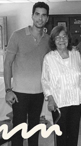 young man in orange red polo standing next to his gramma abuela in a white satin striped shirt and black pants holding a cane