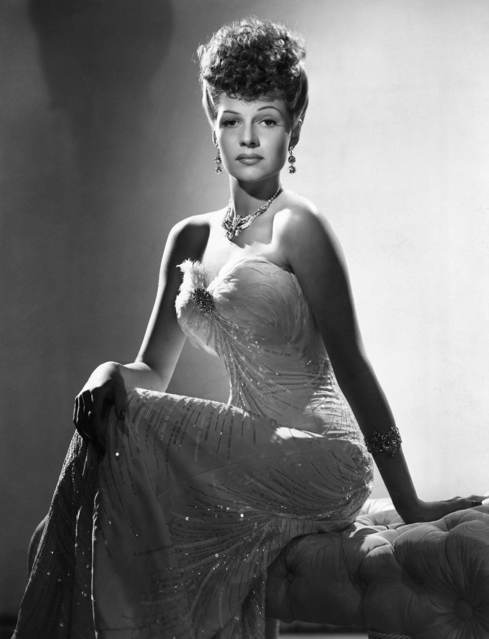 rita hayworth sits on a cushioned stool and looks at the camera, she wears a strapless glittery gown with a necklace and dangling earrings