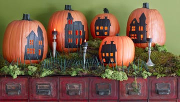 five pumpkins with black houses painted on them and carved out windows, decorated to resemble a village scene