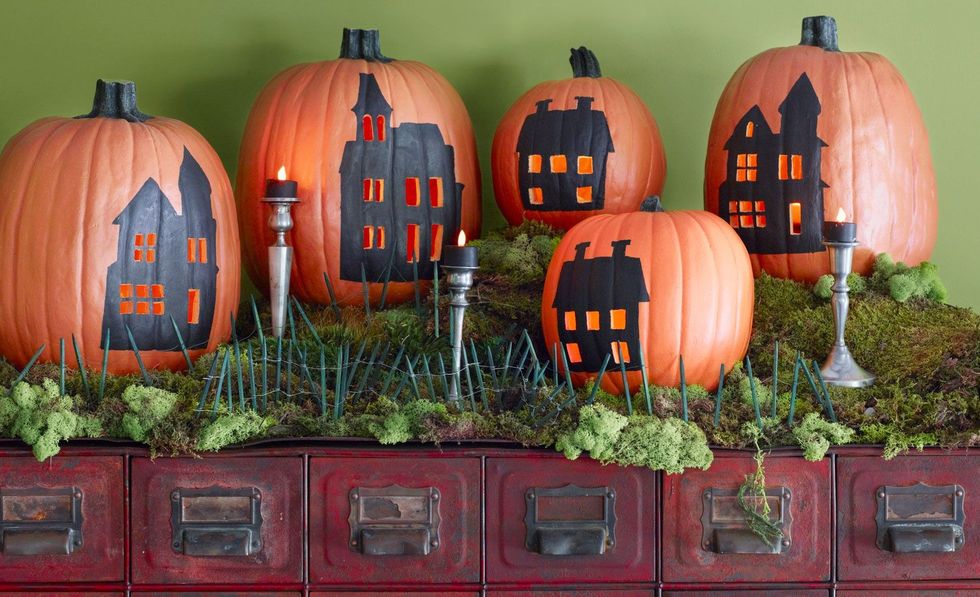 five pumpkins with black houses painted on them and carved out windows, decorated to resemble a village scene