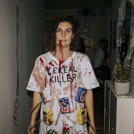 a woman dressed in a gory halloween costume as a cereal killer with fake blood and cereal boxes stabbed with plastic knives glued on to an oversize shirt