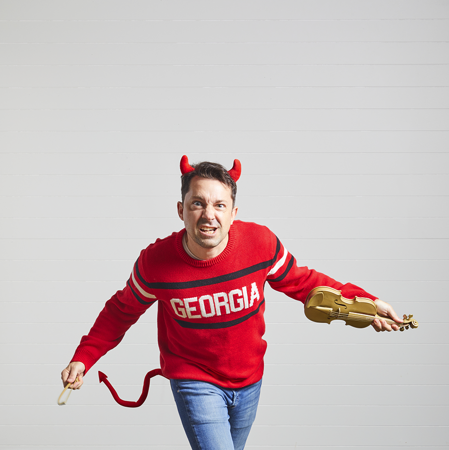 a man dress in a georgia sweater wearing devil horns and tail and has a fiddle painted gold