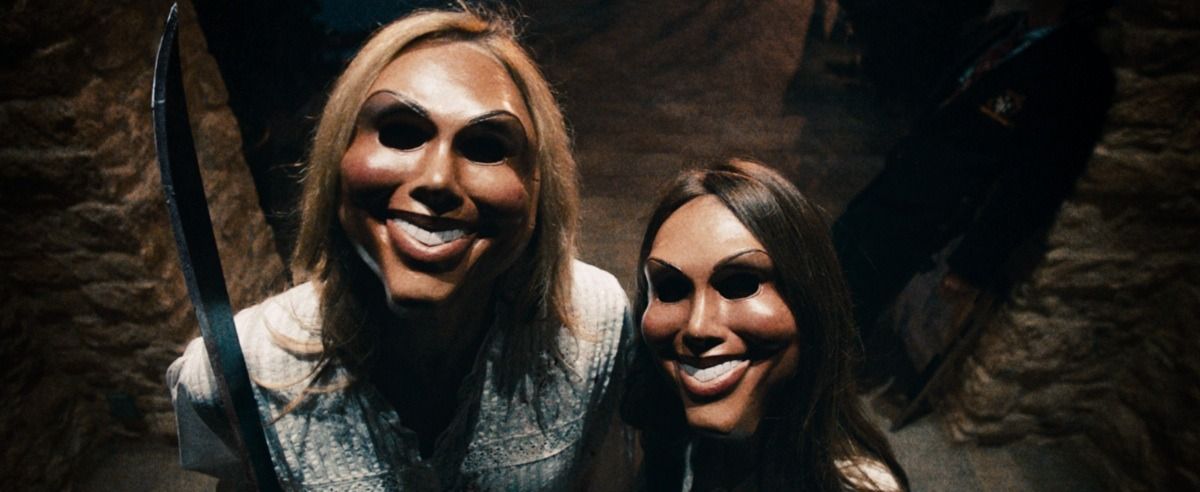two menacing figures stand outside a door while wearing grinning masks in a scene from the purge, the first film if you want to watch the purge movies in order