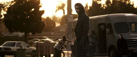 a man wears white face paint with a black, upside down cross painted on his forehead and stitches painted on his lips  in a scene from the purge anarchy, the second film if you want to watch the purge movies in order