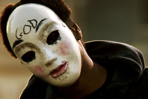 a man wears a white mask with the word god written on it in black in a scene from the purge anarchy