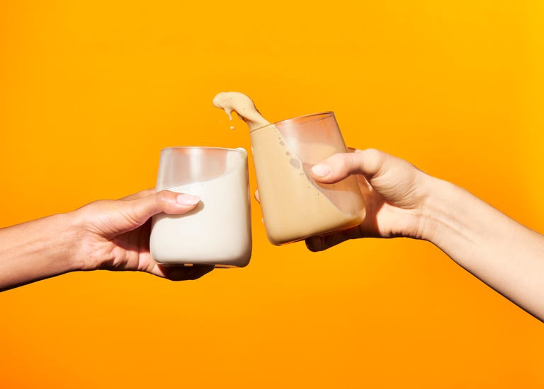 two hands holding milk based drinks clinking glasses in front of an orange background