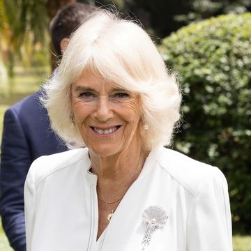 queen camilla smiles at the camera, she wears a white blouse with a pearl and diamond broach along with pearl earrings and simple gold necklaces