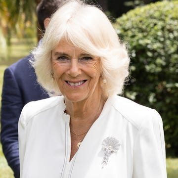 queen camilla smiles at the camera, she wears a white blouse with a pearl and diamond broach along with pearl earrings and simple gold necklaces