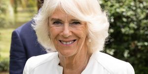 queen camilla smiles at the camera, she wears a white blouse with a pearl and diamond broach along with pearl earrings and simple gold necklaces