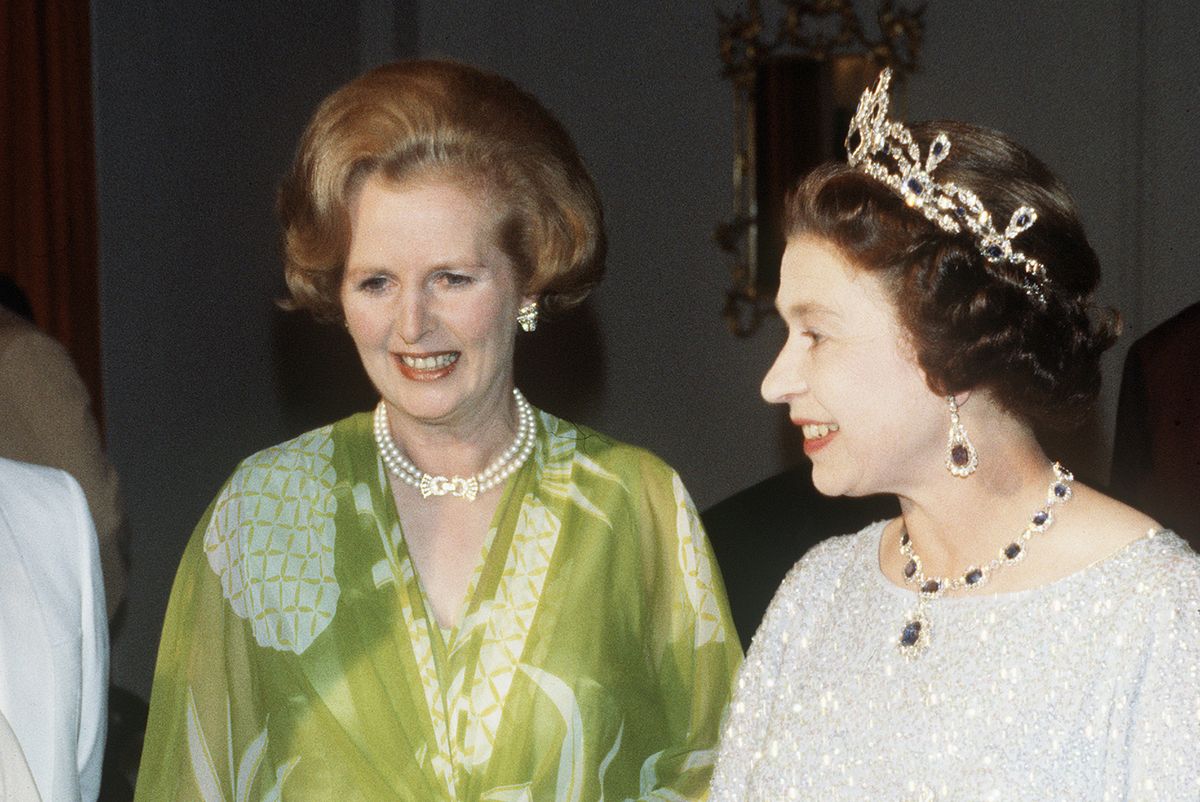 margaret thatcher stands left of queen elizabeth ii at an event during their 1979 trip to zambia, margaret is wearing a lime green dress with a white pineapple pattern, white gloves, a double strand of pearls around her neck and pearl earrings, elizabeth wears a silver sparkly gown, white elbow gloves, and a matching set of diamond and sapphire jewelry, including an opulent pendant necklace, large dangling earrings and a crown, both women are smiling and not looking at the camera