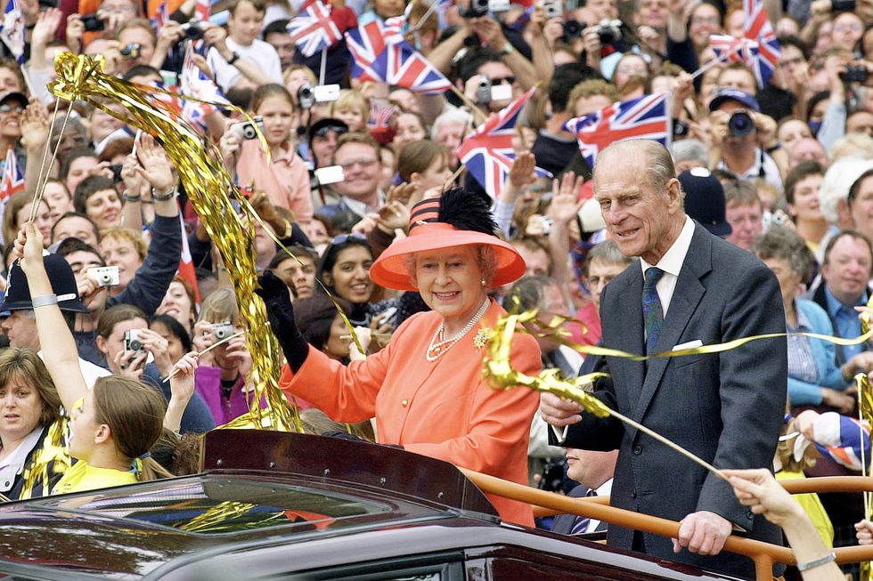 queen elizabeth ii and prince philip stand in the bed of a car that travels through crowds, both smile and wave as people wave british flags and golden streamers, the queen wears an orange outfit and matching hat, the prince wears a gray suit