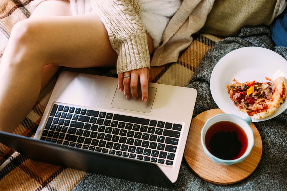 woman's hands visible as she types at laptop sitting in bed with pizza nearby