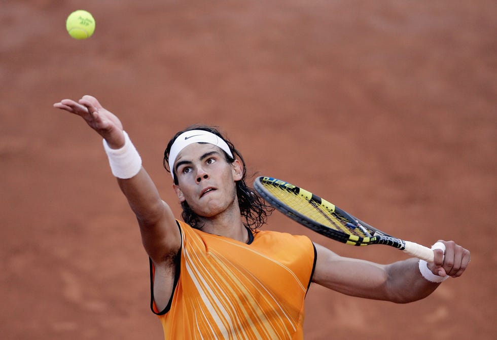 rafael nadal tosses a tennis ball with one outstretched arm as he swings a racket with his other arm, he wears an orange sleeveless tank top, a white headband and white wrist brands