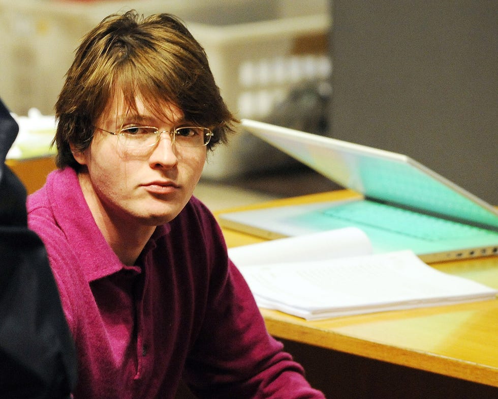a young man sits at a wooden desk and looks to the left, he wears glasses and a pink collared shirt, a computer and papers sit on the desk behind