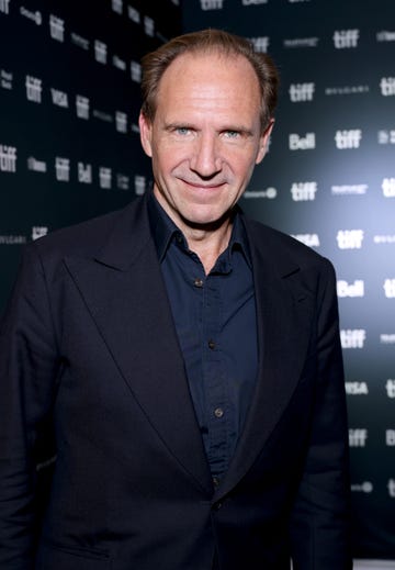 ralph fiennes smiles at the camera, he wears a dark suit jacket and dark navy collared shirt