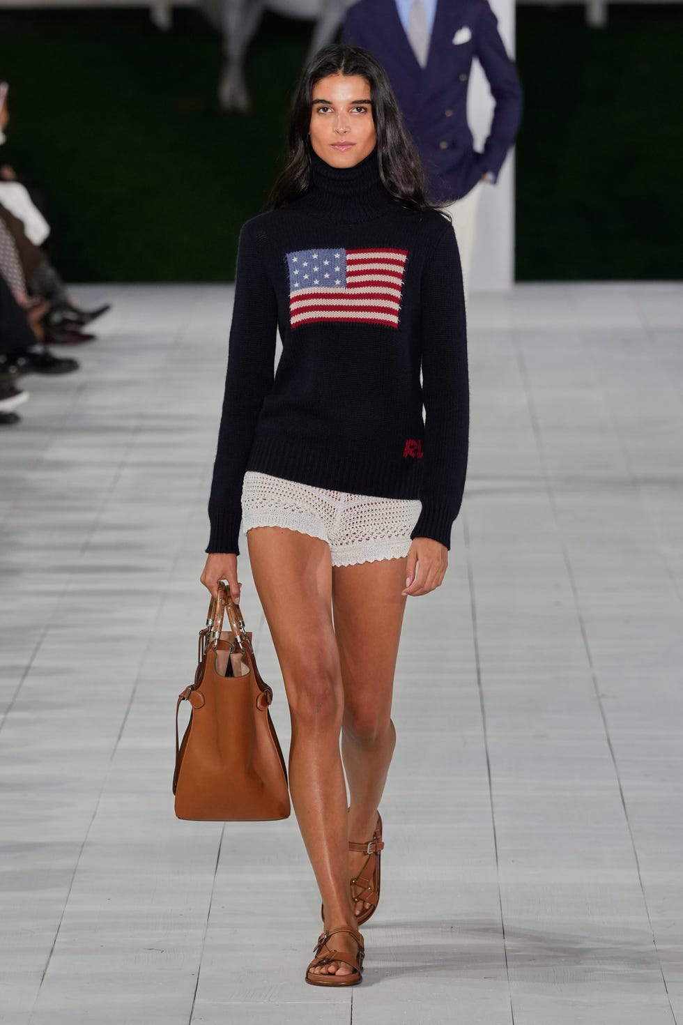 model walking on a runway wearing a sweater with an american flag design and white shorts carrying a brown handbag