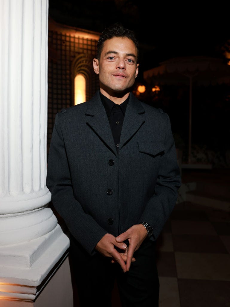 rami malek looks at the camera with his hands clasped in front of him, he wears a dark gray peacoat over a black collared shirt