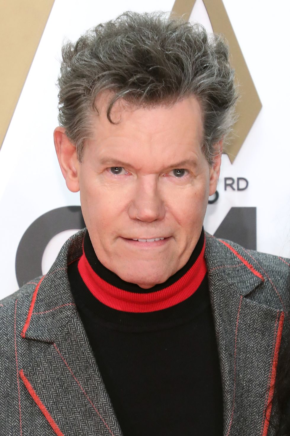 randy travis wearing a black red and gray suit and smiling on an award show red carpet