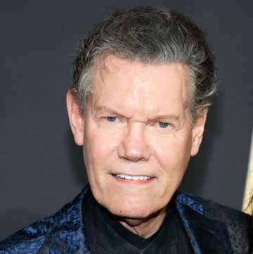 randy travis smiles at the camera, he wears a black shirt and blue and black patterned suit jacket