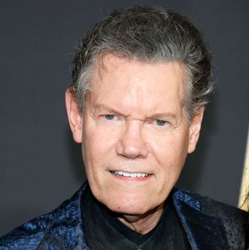 randy travis smiles at the camera, he wears a black shirt and blue and black patterned suit jacket