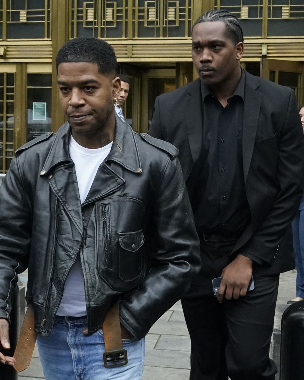 man in black leather jacket walks outside of a concrete building with gold doors, a man follows behind to the right