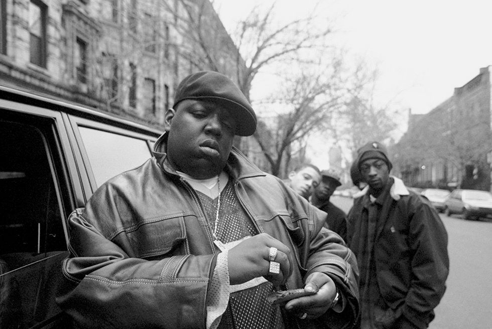 a black and white photo of the notorious big standing next to a car parked on a city street, rolling a cigar, with several people in the background looking at the camera