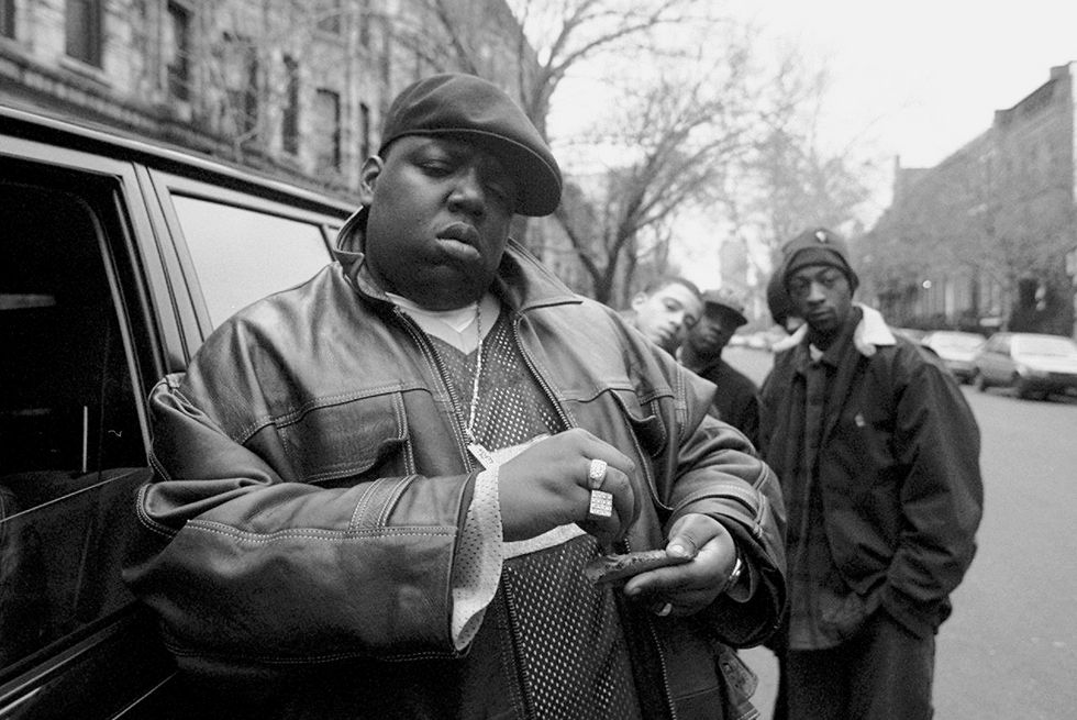 a black and white photo of the notorious big standing next to a car parked on a city street, rolling a cigar, with several people in the background looking at the camera