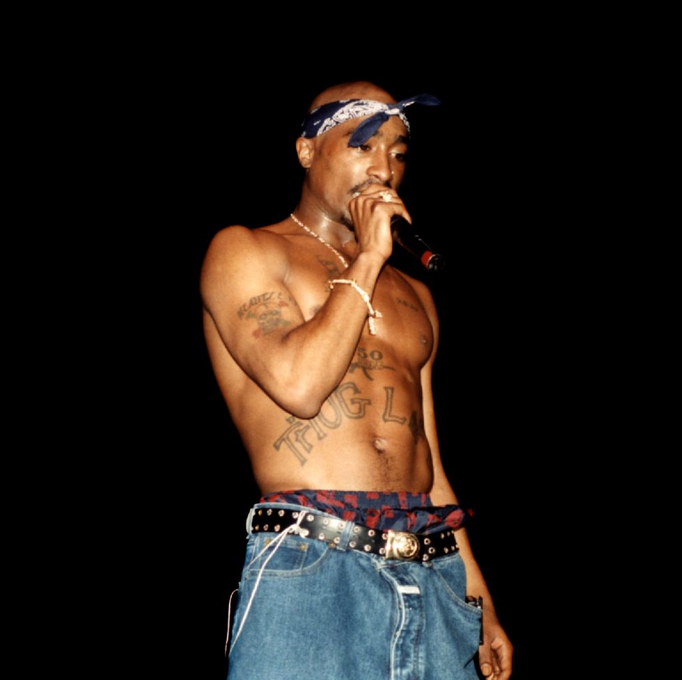 tupac shakur wearing no shirt and jeans, and a bandana on his head, singing into a microphone on a darkened stage