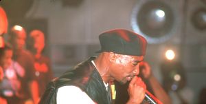 tupac shakur, seen in profile, performs on stage while holding a microphone close to his mouth, he has on a white tshirt, black leather vest, and a backwards black cap