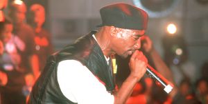 tupac shakur, seen in profile, performs on stage while holding a microphone close to his mouth, he has on a white tshirt, black leather vest, and a backwards black cap