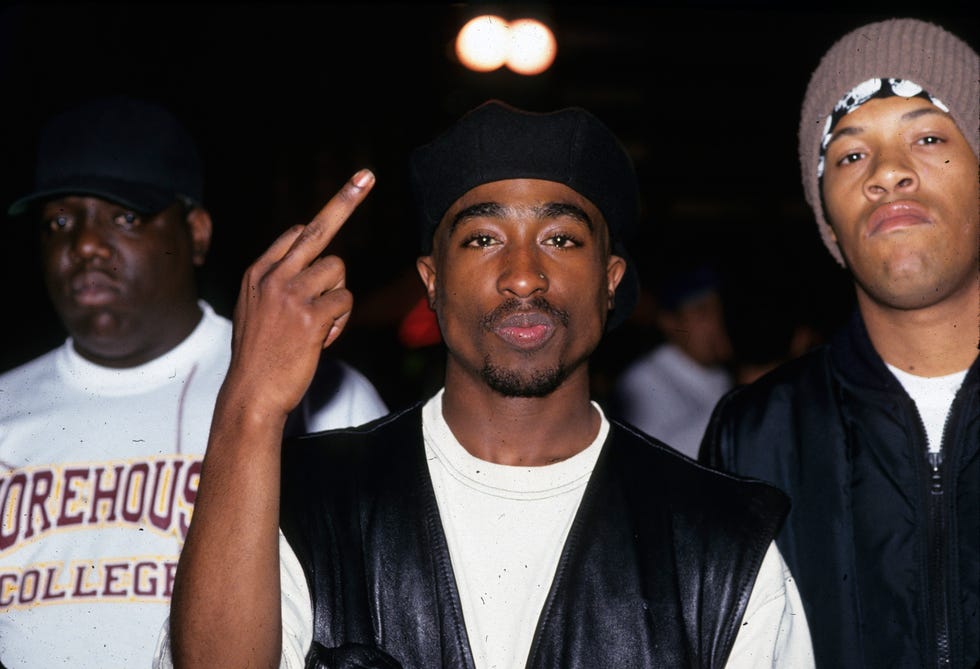 Tupac Shakur in the center wearing a black leather vest and beret, making a defiant gesture with his middle finger raised, flanked by The Notorious B.I.G. in a Morehouse College sweatshirt on the left and another man in a beanie and jacket on the right.