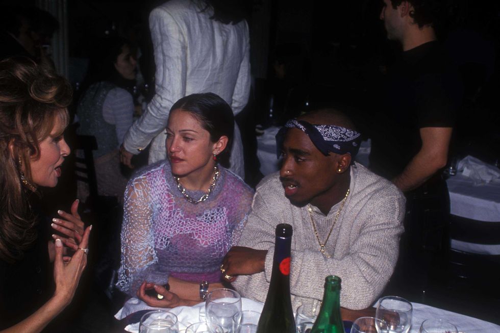 tupac shakur, wearing a white sweater and blue bandana, and madonna, wearing a pink see through shirt, sit at a table with several bottles of alcohol and glasses of water, speaking with raquel welch, who wears a black sleeveless shirt