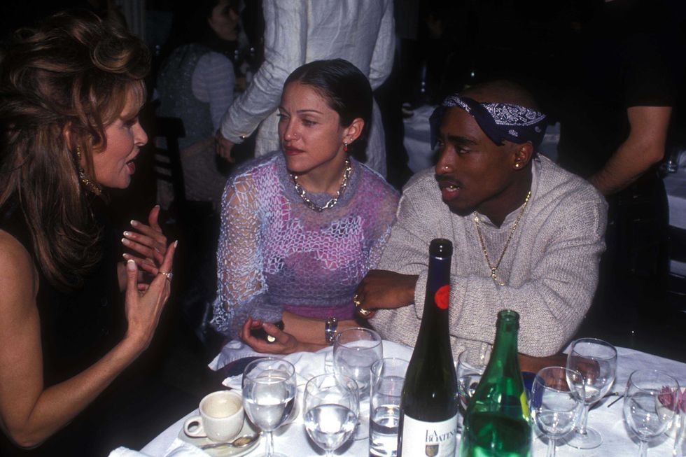 tupac shakur, wearing a white sweater and blue bandana, and madonna, wearing a pink see through shirt, sit at a table with several bottles of alcohol and glasses of water, speaking with raquel welch, who wears a black sleeveless shirt