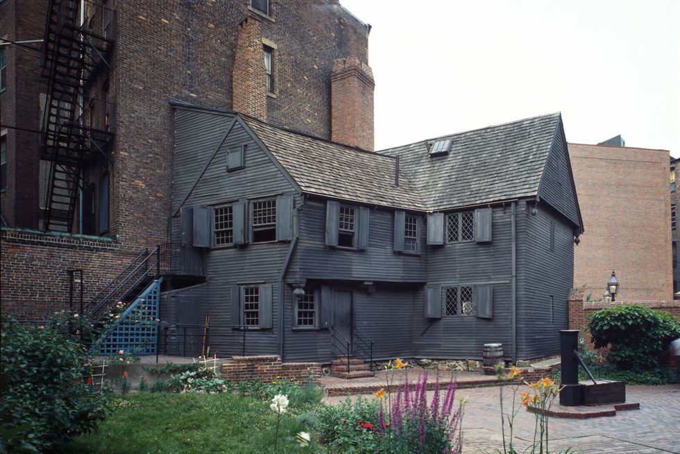 a charcoal color wooden house with many windows stands near brick buildings, flowers and greenery dot the foreground
