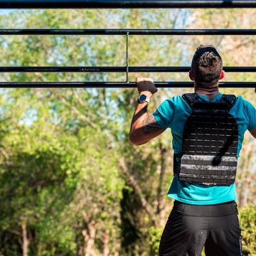 rear view of bearded brunette man doing a barbell pull up with weight vest outdoor fitness concept