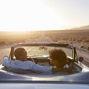 best road trip songs   rear view of couple on road trip driving classic convertible car towards sunset