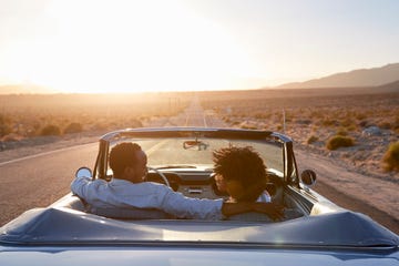 best road trip songs   rear view of couple on road trip driving classic convertible car towards sunset