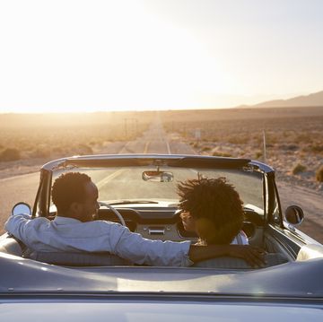 rear view of couple on road trip driving classic convertible car towards sunset