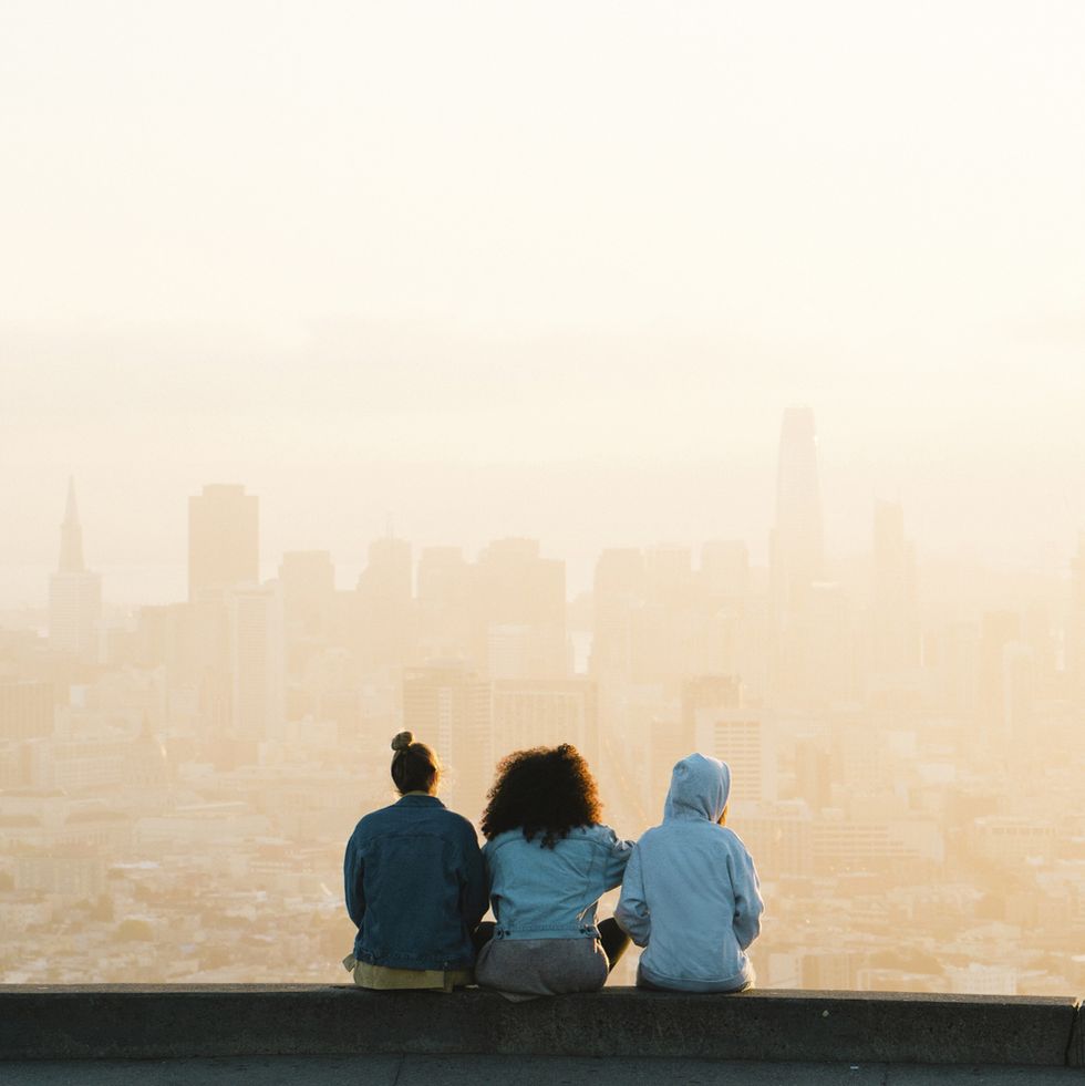 rear view of female friends looking at cityscape while sitting against clear sky during sunset