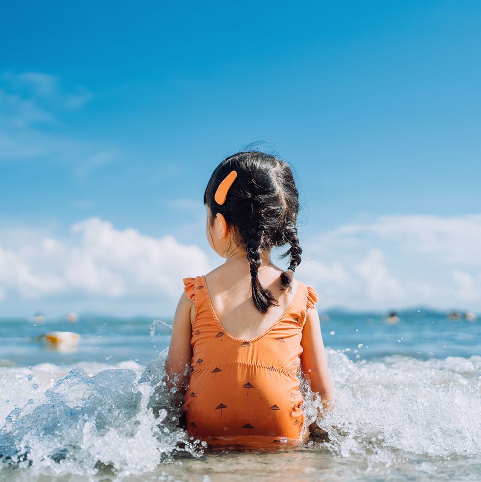 rear view of lovely little asian girl sitting by the seashore at the beach and being splashed by waves. having fun at beach on a sunny summer day