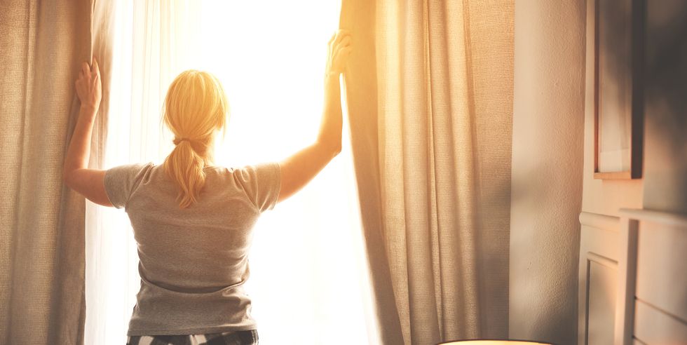 rear view of woman opening the curtains on bright summer morning at sunrise inside her bedroom female opens drapes to let the sunlight in her room lady wearing pajamas awake and ready for the day