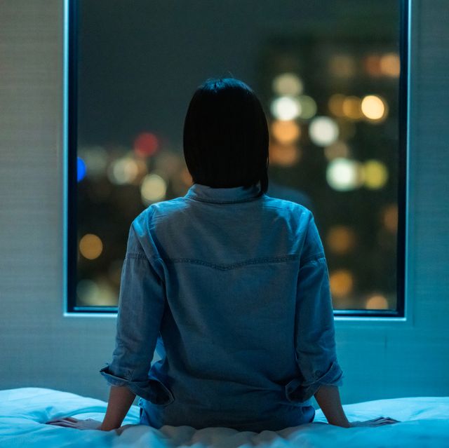 rear view of woman sitting alone on bed in room and looking through window at night
