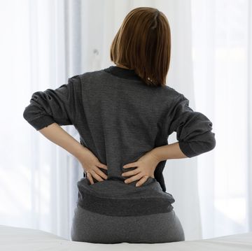rear view of woman touching back in pain while sitting on bed at home