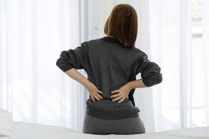 rear view of woman touching back in pain while sitting on bed at home