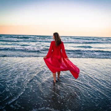 rear view of woman wearing red dress walking on shore at beach against sky during sunset
