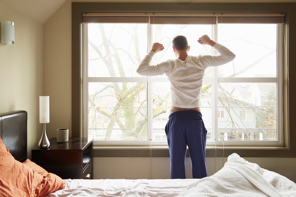 Rear view of young man stretching in front of bedroom window