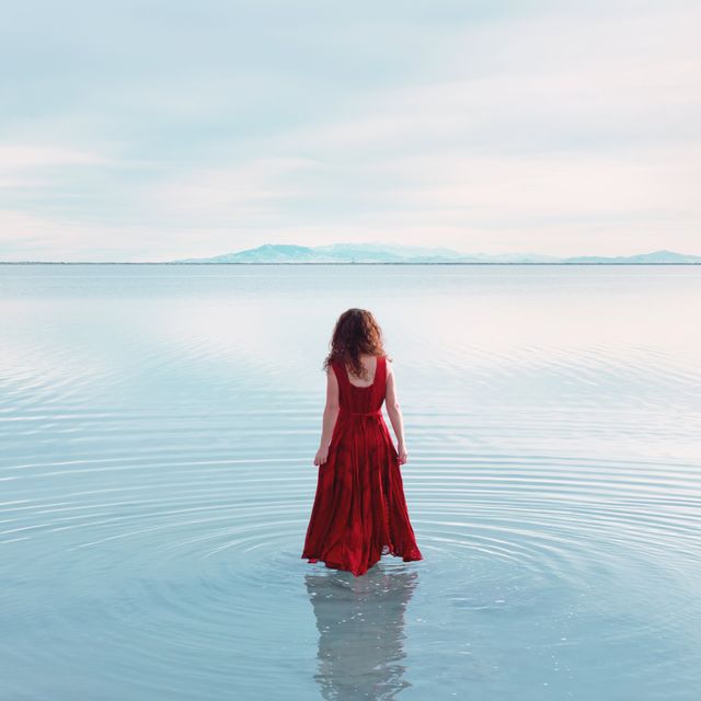rear view of young woman with long wavy red hair in sleeveless red dress walking in shallow still lake