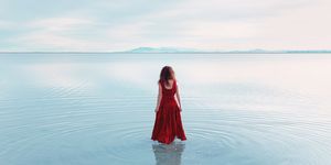 rear view of young woman with long wavy red hair in sleeveless red dress walking in shallow still lake