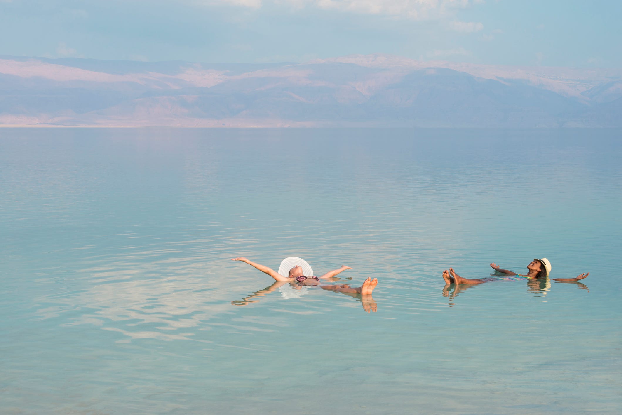 lovely multiracial girls wearing sun hats, lying on back with outstretched arms, floating in salty water of dead sea unusual buoyancy caused by high salinity